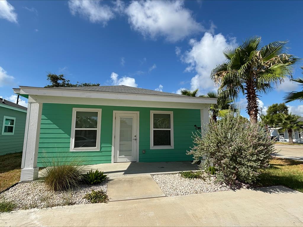 1-bedroom, 1-bath house in Oasis Resort community, featuring green exterior, landscaped front yard with palm trees and shrubs, and clear blue sky.